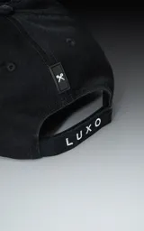 Gorra negra de cinco paneles con visera plana y un pequeño parche cuadrado blanco en el frente con un logo bordado en forma de tijeras cruzadas.