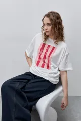 Remera blanca de calce relajado y silueta amplia, con cuello redondo, mangas cortas y estampa gráfica frontal de un cangrejo sobre franjas en tonos rosado y rojo.