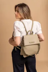 Mochila tipo cartera con tapa, confeccionada en símil cuero color suela. Cuenta con doble compartimento, división interna, cierre de seguridad y bolsillo trasero con cierre. Incluye una correa frontal decorativa con tachas metálicas.
