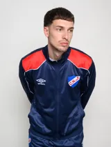 Conjunto deportivo Umbro del Club Nacional de Football, compuesto por una campera con cierre frontal y pantalón a juego. La campera es de color azul con detalles en rojo en los hombros y líneas blancas, presentando el logo de la marca y el escudo del club en el pecho. El conjunto está confeccionado en tejido sintético con acabado satinado.