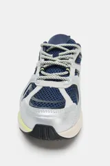 Zapatilla deportiva estilo running con combinación de materiales y colores. Presenta paneles en azul marino, plateado y detalles en amarillo neón, con suela de plataforma en tono beige y tirador en el talón.