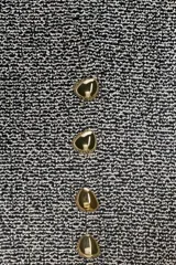 Chaleco de tejido texturado en tonos claros (blanco/beige/gris) con escote pico y cierre frontal de cuatro botones dorados. Presenta un bajo asimétrico.