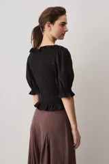Blusa negra con cuerpo fruncido tipo smock, mangas cortas abullonadas con puños elásticos y volados en el cuello y el ruedo.