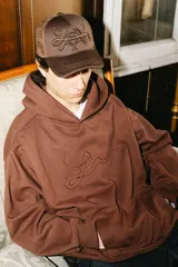 Gorra trucker marrón con frente de tela y parte trasera de rejilla. Presenta un bordado 3D con un diseño abstracto en el frente.