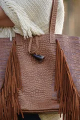 Bolso tipo shopper de cuero con textura de cocodrilo, color marrón. Presenta flecos laterales largos, correas de hombro y un detalle decorativo de guampa colgante.