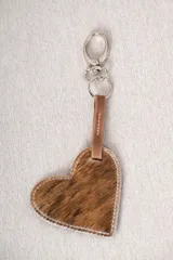 Llavero con dije en forma de corazón confeccionado en cuero con pelo, con costuras blancas a la vista y tira de sujeción en cuero liso, unido a una argolla y mosquetón metálicos.