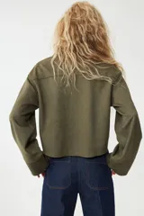 Camisa corta color verde militar, de corte holgado, con cuello clásico, botones frontales y canesú en la espalda. Presenta hombros caídos y mangas largas con puños anchos.