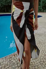 Falda midi de corte cruzado con diseño tipo pareo, presenta un nudo decorativo en la parte delantera y cierre de cremallera oculta en la espalda. El tejido cuenta con un estampado geométrico abstracto en tonos bordó, negro, amarillo y crema.