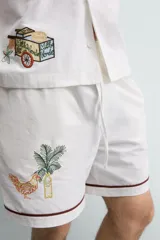 Bermuda de corte relajado confeccionada en algodón, color blanco con vivo en contraste color marrón en el ruedo. Presenta cintura elástica con cordón ajustable y un bordado decorativo en la pierna derecha que incluye una palmera y un gallo.