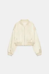 Chaqueta bomber corta de tejido satinado color beige, con cuello redondo, cierre frontal con cremallera, mangas largas de hombro caído y bolsillos de vivo en el delantero. Presenta acabados en rib elástico en cuello, puños y ruedo.