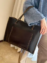 Cartera de cuero genuino color chocolate, tipo tote bag, con asas trenzadas, costuras artesanales visibles en los bordes y un amplio bolsillo frontal.
