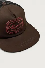 Gorra trucker marrón con visera plana y red negra en la parte trasera. Cuenta con un bordado rojo en el frente con la inscripción "Indie Behaviour 1975" y otro bordado blanco en el lateral que dice "Committed to the bands".
