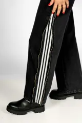 Pantalón de jean negro con corte recto, tiro alto, bolsillos delanteros y traseros, cierre con cremallera y botón. Presenta tres líneas blancas verticales a lo largo de la pierna y cierres metálicos dorados.