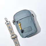 Bolso porta celular celeste de tela con cierre superior, correa larga estampada ajustable y charm decorativo.