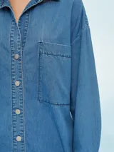 Camisa de denim azul de corte oversize, con mangas largas, cuello clásico, un bolsillo aplicado en el pecho y cierre frontal con botones metálicos.