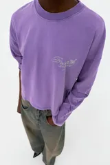 Camiseta de manga larga con corte oversize, confeccionada en algodón color violeta con efecto lavado. Presenta cuello redondo, bajo asimétrico y detalles de texto con aplicación de brillos en el pecho y las mangas.