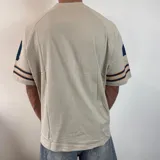 Remera color crema de corte oversize, con cuello en V y mangas cortas. Estampado frontal con el número 43 en azul, flores naranjas y gaviotas blancas. Tiene rayas azules y naranjas en las mangas.