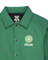 Chaqueta tipo coach verde con cuello camisero, cierre frontal de botones a presión y cordón ajustable en el ruedo. Presenta un logo circular blanco con texto en el pecho izquierdo.