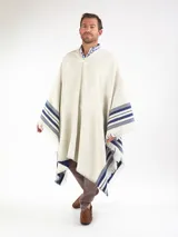 Poncho grueso color beige con detalles de rayas azules en los laterales y flecos en los bordes. Presenta el escudo bordado en la espalda y diseño liso en la parte frontal con escote en V.
