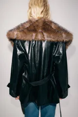 Trench corto Zara de efecto piel color negro, con cuello de solapa y aplicación de pelo sintético a contraste en color marrón. Cuenta con manga larga con trabillas en los puños, bolsillos delanteros y cinturón ajustable del mismo tejido.