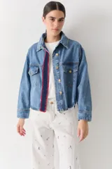 Campera de jean de corte cropped, con cierre frontal de botones metálicos. Presenta un detalle decorativo tejido a mano en crochet con bordes rojos en la solapa y un parche lateral. El diseño incluye bolsillos frontales con solapa y un bordado en la parte posterior, con un corte asimétrico más largo en la espalda.