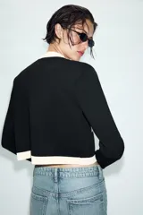 Chaqueta de punto de corte recto y manga larga, con cuello redondo. Presenta un diseño bicolor en negro y crema, con cierre frontal de botones dorados y bolsillos de plastrón en el delantero con detalles en contraste.