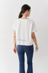 Remera blanca de algodón con cuello redondo, manga corta y bordes con detalle de costura en hilo celeste. Presenta un bordado central de un sol amarillo y la palabra 'SAVIA' debajo.