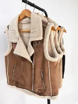 Chaleco estilo gamulán de gamuza color marrón con interior y bordes de corderito sintético color crema. Presenta cierre frontal metálico, bolsillos laterales con cierre y detalles de tiras de cuero sintético en la cintura.