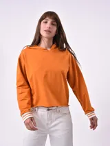 Hoodie de color naranja con capucha, manga larga y puños acanalados con rayas blancas. Presenta un corte recto y holgado.