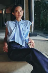 Camisa sin mangas confeccionada en algodón, con cuello redondo y detalle de lazada frontal. Presenta un diseño de corte holgado con bajo redondeado y cierre posterior mediante abertura y botones.