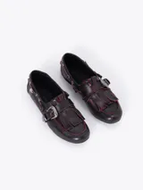 Zapato tipo mocasín de cuero vacuno con efecto gastado y detalles en color rojizo. Presenta flecos en la capellada, una hebilla metálica decorativa y herrajes plateados a lo largo del lateral.