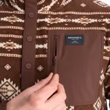 Buzo polar marrón con estampado geométrico en tonos beige y blanco, cuello alto con cierre de botones a presión, bolsillo tipo canguro y bolsillo en el pecho con cierre. Marca Merrell.