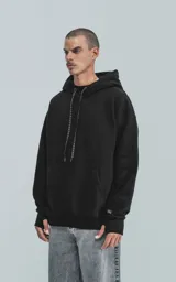 Hoodie negro de frisa premium, corte oversize, con capucha ajustable y puños y cintura elastizados. Presenta un bolsillo canguro frontal y un detalle de etiqueta tejida con texto repetido en los cordones de la capucha y en la manga izquierda.