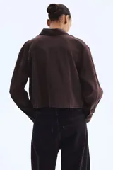 Camisa marrón de manga larga, corte corto y diseño holgado, con cuello clásico, cierre frontal de botones, bolsillos con solapa en el pecho y cordón ajustable en el bajo.