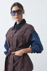 Chaleco de corte relajado con estampado a cuadros en tonos tierra, cuello camisero oscuro y cierre frontal con botones. Incluye un cinturón de la misma tela para ajustar a la cintura y bolsillos frontales.