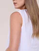 Blusa blanca sin mangas, confeccionada en algodón con sutiles rayas verticales en relieve. Presenta cuello redondo, cierre frontal con botones de madera y un nudo ajustable en el dobladillo delantero.