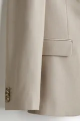 Blazer color beige de corte slim fit, con solapas de muesca finas, ojal decorativo, botonadura sencilla, bolsillo superior insertado, bolsillos delanteros con solapa, dos bolsillos interiores, botones decorativos en los puños y abertura doble en la parte posterior.