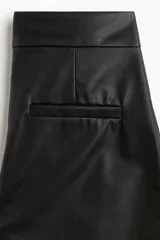 Pantalón de cuero sintético negro con corte bootcut, cierre frontal con botón oculto y costuras decorativas sobre las rodillas.