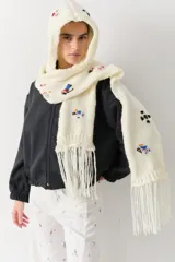 Bufanda con capucha integrada, confeccionada en tejido de punto grueso color crudo. Presenta bordados coloridos de figuras deportivas y cuenta con flecos en los extremos.