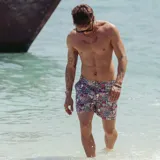 Short de baño con estampado floral y botánico en tonos violeta, rosa y verde sobre fondo oscuro. Presenta pretina fija con hebillas metálicas laterales para ajuste, cierre frontal con botón a presión, bolsillos laterales y bolsillo trasero.