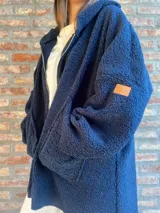 Campera de corderito color verde oscuro, con diseño holgado, cuello alto y manga larga. Presenta una etiqueta de cuero sintético en la manga.