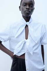 Blusa a rayas celestes y blancas, de manga corta abullonada, con cuello adornado con lazos y cierre frontal con botones.