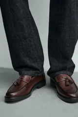 Mocasín de vestir color marrón oscuro, con parte superior lisa, detalle de antifaz con borlas en el empeine y horma redonda. Suela de goma a tono.