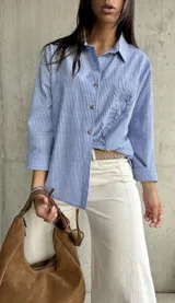 Camisa clásica de manga larga, a rayas verticales azules y blancas, con cuello camisero y botones frontales. Presenta un bordado lineal de un rostro en el frente.