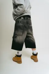 Bermuda de jean estilo carpintero, corte baggy, color negro lavado con efecto degradado. Presenta bordados en la parte trasera con tipografía gótica que dice "Lucca Bossio". Incluye bolsillos grandes y bolsillo lateral de carpintero.