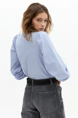 Camisa de algodón a rayas verticales celestes y blancas, con cuello mao, mangas largas abullonadas con puños elásticos y detalle de volado en la cartera de botones frontal.