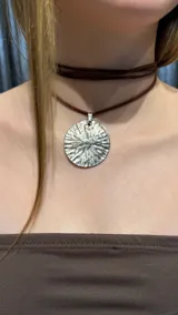 Gargantilla tipo choker compuesta por una tira de gamuza marrón que rodea el cuello, con un dije circular de metal plateado con textura irregular y acabado brillante.