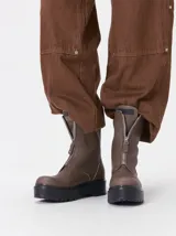 Botas de cuero color marrón de caña alta, con plataforma gruesa de goma y cierre metálico frontal con tirador. Presentan costuras a contratono y detalle de etiqueta de la marca en la lengüeta.