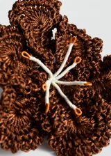 Broche en forma de flor de gran tamaño, confeccionado en tejido de crochet color marrón, con detalle central de pistilos en hilo blanco y naranja.