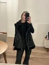 Chaleco negro con cuello solapa y borde de piel sintética, cierre frontal con cremallera y bolsillos laterales con cremallera.